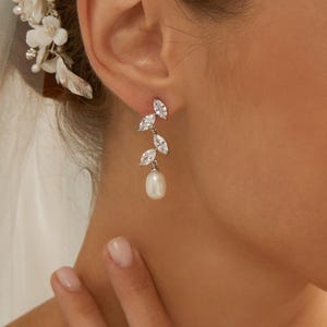 May include: Elegant silver earrings with a leaf design, featuring sparkling clear crystals and a dangling white pearl. The earrings are designed for a formal occasion, such as a wedding or prom.