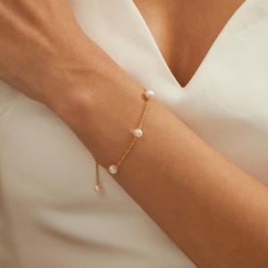 May include: A delicate gold bracelet featuring several small, round, white pearls. The bracelet is attached to a fine gold chain. The jewelry is worn on a person's arm, against a white background.