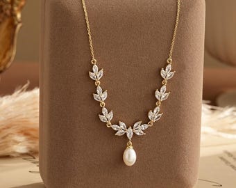 Puede incluir: Un collar de tono dorado con una cadena delicada adornada con cristales transparentes de corte marquesa dispuestos en un patrón de hojas, centrado con un colgante de perla blanca en forma de lágrima que cuelga de la base del diseño.