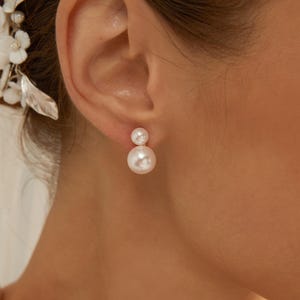 Op de afbeelding: Een paar elegante, witte parel oorbellen. Elke oorbel heeft een kleine parel boven een grotere parel, wat een klassiek ontwerp creëert. De oorbellen worden op een oor getoond.