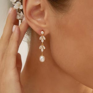 May include: A gold drop earring featuring two marquise-cut clear stones arranged in a leaf-like pattern, suspended from a small round stud, and finished with a single white teardrop pearl dangling at the bottom of the delicate metal chain.