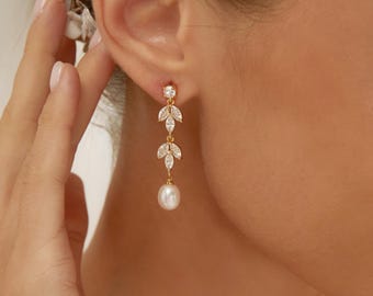 Puede incluir: Un pendiente colgante dorado con dos piedras transparentes de talla marquesa dispuestas en forma de hoja, suspendidas de un pequeño perno redondo y rematado con una perla blanca en forma de lágrima que cuelga de la fina cadena metálica.