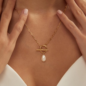 May include: A gold necklace featuring a toggle clasp and a pear-shaped pearl pendant. The chain is a delicate link style. The necklace is worn against a white top.