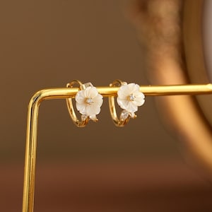 May include: A pair of gold-colored hoop earrings, each adorned with a white flower design. The flowers have a small, clear gem in the center and are accented with additional clear gems. The earrings are displayed on a gold-colored stand.