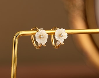 以下が含まれることがあります： 中央に小さな透明な石をあしらった白い彫刻の花のチャームと、小さな葉の形のアクセントが付いたゴールドのフープピアス。ピアスはゴールドトーンの金属製スタンドに飾られており、繊細な花のデザインとメタリックな仕上げが特徴です。