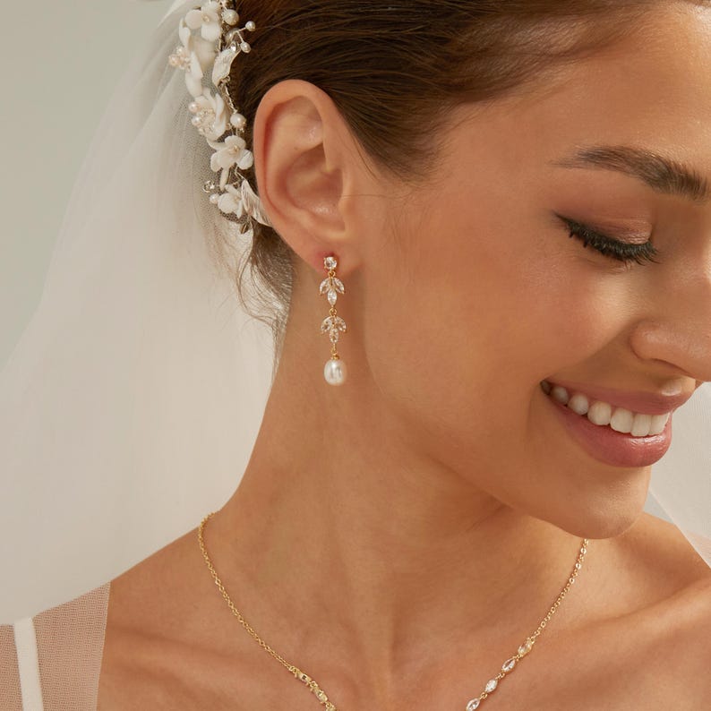 May include: A gold-toned drop earring featuring two marquise-cut clear crystals arranged in a leaf-like pattern, finished with a single white pearl at the base. The earring is worn alongside a matching gold chain necklace with small clear crystals.