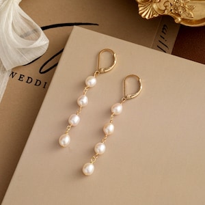 Può includere: Un paio di eleganti orecchini pendenti dorati con una serie di perle bianche. Gli orecchini hanno una chiusura a leva e sono esposti su una superficie dai toni neutri, suggerendo un tema di gioielli o matrimonio.