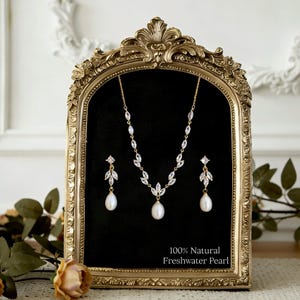 Puede incluir: Un conjunto de joyería en tono dorado que incluye un collar y dos pendientes. Cada pieza presenta piedras transparentes de talla marquesa en patrones de hojas y una perla de agua dulce blanca. El texto 100% Natural Freshwater Pearl es visible.