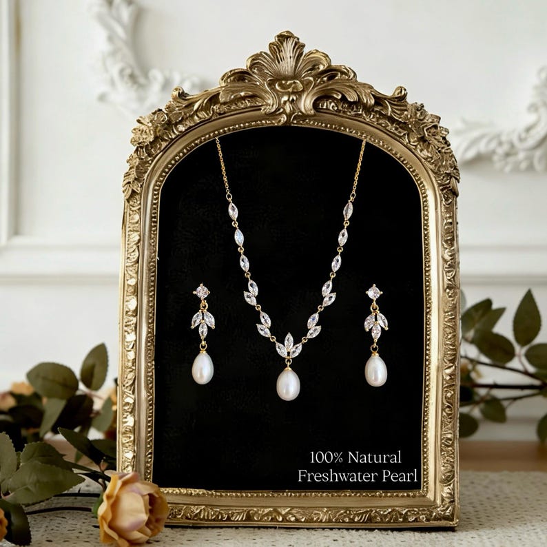 May include: A gold-toned jewelry set featuring a necklace and two drop earrings. Each piece includes marquise-cut clear stones arranged in leaf-like patterns and a single white freshwater pearl. The text 100% Natural Freshwater Pearl is visible.