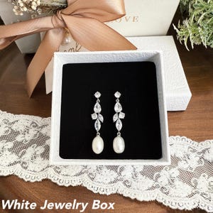 May include: A pair of silver-toned drop earrings featuring clear, faceted marquise and pear-shaped crystals arranged in a vertical cluster, terminating in a smooth, white teardrop-shaped pearl, presented inside a square white jewelry box.