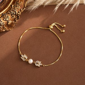 May include: Gold-toned bracelet with a delicate chain and a sliding clasp. The bracelet features a central pearl and two leaf-shaped accents adorned with clear crystals. The design is elegant and suitable for various occasions.