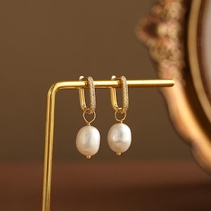 Puede incluir: Un par de pendientes de aro dorados con una perla blanca colgante. Los aros están incrustados con pequeñas piedras transparentes. Los pendientes se muestran sobre un soporte dorado.