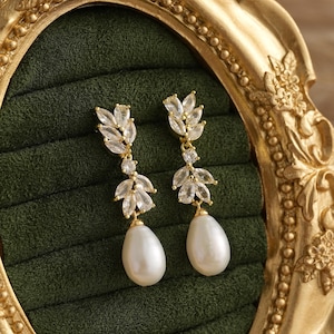 Peut inclure: Une paire de boucles d'oreilles pendantes dorées. Chaque boucle d'oreille présente un motif en forme de feuille avec des cristaux transparents et une grosse perle blanche en forme de poire. Les boucles d'oreilles sont présentées sur un fond en velours vert foncé.