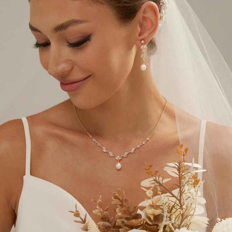 May include: A gold-toned necklace and matching drop earrings set featuring clear marquise-cut crystals arranged in a leaf-like pattern. The necklace features a central teardrop-shaped white pearl pendant, while each earring has a single pearl drop.