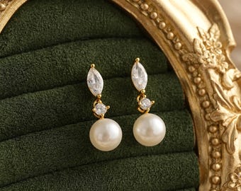 May include: A pair of gold-tone drop earrings featuring a marquise-cut clear stone at the top, a small round clear stone in the center, and a round white pearl at the bottom. The earrings are displayed against a dark green velvet-lined surface.