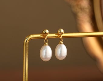 May include: A pair of gold-colored drop earrings featuring lustrous, white, oval-shaped pearls. The earrings are suspended from small, round gold studs. The pearls have a smooth, polished surface, reflecting light. The earrings are displayed on a gold-colored stand.