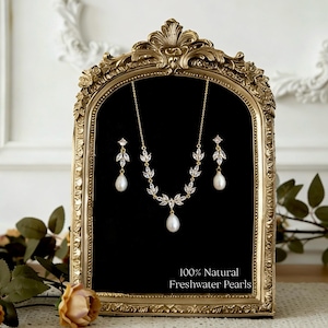 Może przedstawiać: Złoty naszyjnik i kolczyki z perłami słodkowodnymi i akcentami w kształcie liści. Biżuteria jest prezentowana na czarnym tle w złotej, ozdobnej ramie. Widoczny jest tekst "100% Natural Freshwater Pearls".