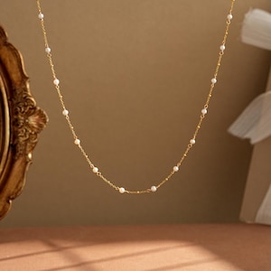 Op de afbeelding: Een delicate gouden ketting met kleine, gelijkmatig verdeelde parels. De ketting heeft een fijne ketting en een eenvoudig, elegant ontwerp. De parels zijn wit en de ketting heeft een warme goudkleur. De ketting wordt gepresenteerd tegen een neutrale achtergrond.