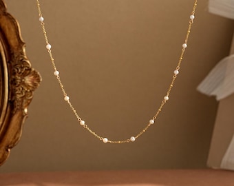 May include: A delicate gold necklace featuring small, evenly spaced pearls. The necklace has a fine chain and a simple, elegant design. The pearls are white and the chain is a warm gold color. The necklace is displayed against a neutral background.