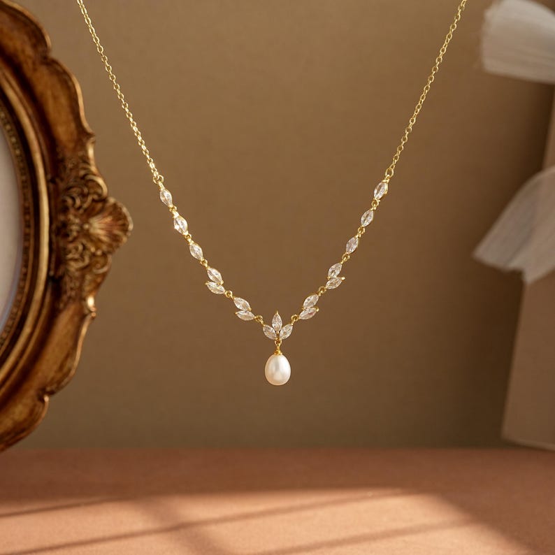 May include: A gold chain necklace featuring a series of marquise-cut clear crystals arranged in a leaf-like pattern along the sides, culminating in a central drop pendant with a single white pearl suspended from the bottom of the crystal cluster.