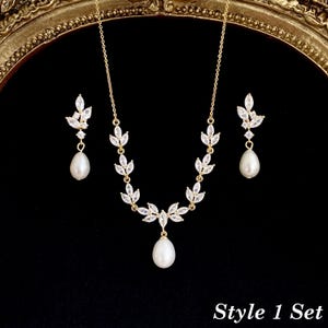 May include: A gold-toned jewelry set consisting of a necklace and two earrings. The necklace features a chain with marquise-cut clear stones arranged in a leaf pattern, ending in a central white teardrop pearl. Matching earrings feature a leaf-shaped stone cluster with a dangling white pearl. Text reads: Style 1 Set.