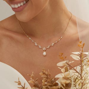 May include: A gold-toned necklace featuring a delicate chain adorned with marquise-cut clear stones arranged in a leaf-like pattern, centered with a single teardrop-shaped white pearl pendant that hangs from the middle of the decorative arrangement.