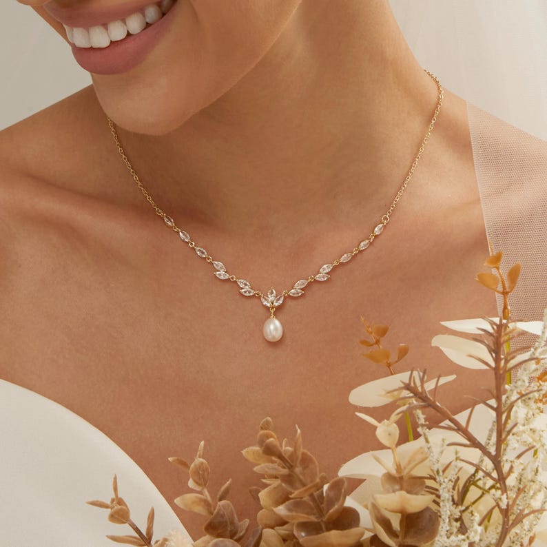 May include: A gold-toned necklace featuring a delicate chain adorned with marquise-cut clear stones arranged in a leaf-like pattern, centered with a single teardrop-shaped white pearl pendant that hangs from the middle of the decorative arrangement.