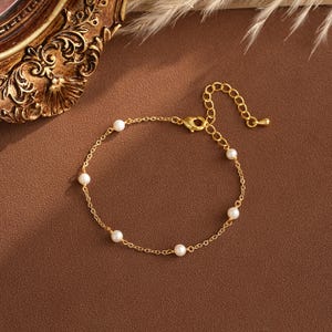 May include: A delicate gold-toned bracelet featuring small, round, white pearls. The bracelet has a chain-link design with an adjustable clasp. The pearls are evenly spaced along the chain, creating a simple yet elegant look. The background is a warm brown.