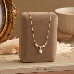 May include: A gold-colored necklace with a floral design and a single pearl pendant. The necklace is displayed on a brown velvet jewelry box. The text "Rayon de soleil" is visible on the book below the box.