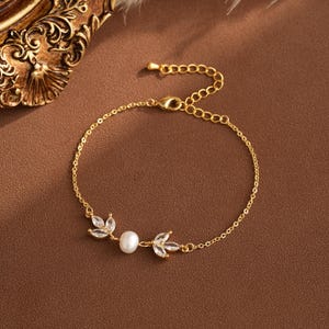 May include: Gold-toned bracelet featuring a delicate leaf design with clear crystals and a central pearl. The adjustable chain adds versatility. The bracelet is displayed on a brown surface.