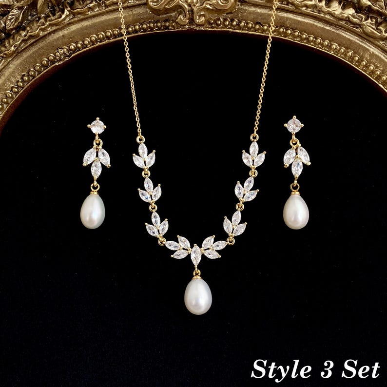 May include: A gold-toned jewelry set consisting of a necklace and two matching earrings. The necklace features a chain with marquise-cut clear stones arranged in leaf-like patterns, ending in a single white teardrop pearl pendant. The earrings mirror this design with a small stud, leaf-shaped stones, and a dangling white pearl. The text "Style 3 Set" is visible in the bottom right corner.