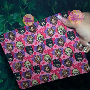 May include: A pink rectangular item featuring a repeating pattern of Bratz doll faces within purple hearts and red lips. The word "Bratz" is printed in purple. The item is held against a green leafy background.