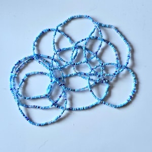 May include: Several blue beaded necklaces in shades of light and dark blue. The small beads are strung together, forming a delicate design. The necklaces are arranged in a tangled, circular pattern. The beads are measured in centimetres.