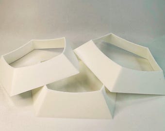 May include: Four white, geometric-shaped objects with a wide base and angled sides. The objects appear to be made of a solid material and are arranged on a white surface. The shapes have a collar-like appearance.
