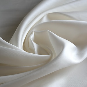 Pode incluir: Uma peça de tecido na cor branco-sujo com uma superfície lisa e lustrosa e um padrão de trama diagonal fino. O material está drapeado em dobras suaves e fluidas que criam sombras e reflexos profundos, enfatizando o brilho do tecido.