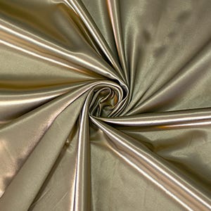 Può includere: Un pezzo di tessuto color oro metallizzato con una superficie liscia e riflettente. Il materiale è raccolto in una spirale centrale, creando pieghe profonde e fluide con riflessi che enfatizzano la lucentezza satinata del tessuto.