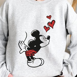 K&ouml;nnte beinhalten: Ein hellgraues Sweatshirt mit einer Grafik von Micky Maus, die K&uuml;sse pustet, mit drei roten Herzen, die dar&uuml;ber schweben. Micky ist in seiner klassischen schwarzen, roten und wei&szlig;en Kleidung dargestellt. Das Sweatshirt hat lange &Auml;rmel und einen Rundhalsausschnitt.