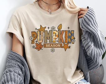 May include: Beige t-shirt with the words "PUMPKIN SEASON" in a fall-themed design. The text features orange, brown, and blue patterns, with leaf and star accents. The shirt is a casual, short-sleeved style.