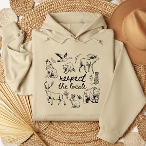 Puede incluir: Sudadera con capucha beige con un gr&aacute;fico negro de varios animales, incluyendo un lobo, un alce y un oso, con el texto "respect the locals". La sudadera tiene una capucha con cord&oacute;n y mangas largas.