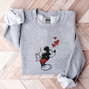 K&ouml;nnte beinhalten: Hellgraues Sweatshirt mit einer Grafik von Mickey Mouse, die drei rote Herzen pustet. Mickey ist in Schwarz, Rot und Wei&szlig; dargestellt. Das Sweatshirt hat einen Rundhalsausschnitt und lange &Auml;rmel.