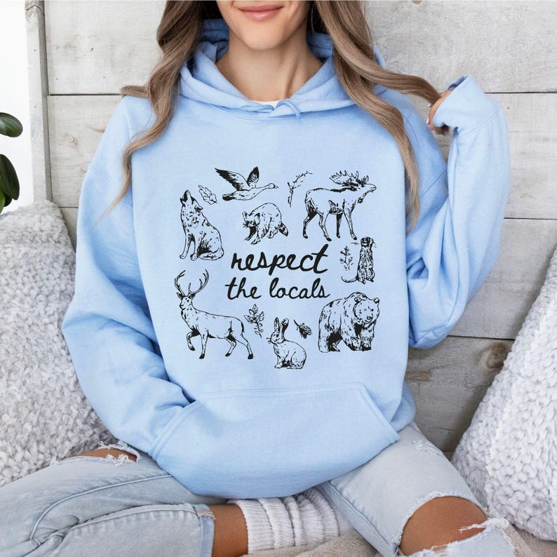 Puede incluir: Sudadera con capucha azul claro con un gr&aacute;fico negro de varios animales del bosque y el texto "respect the locals". El dise&ntilde;o incluye un lobo, un oso, un ciervo, un conejo y otros animales. La sudadera tiene un bolsillo delantero y una capucha.