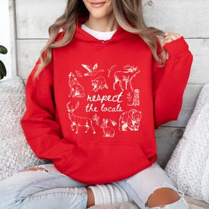 Puede incluir: Una sudadera con capucha roja con un gr&aacute;fico blanco de varios animales, incluyendo un lobo, un oso y un ciervo. El texto "respect the locals" est&aacute; impreso debajo de las ilustraciones de animales. La sudadera est&aacute; hecha de un material suave.