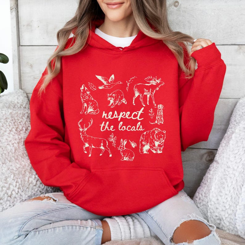 Puede incluir: Una sudadera con capucha roja con un gr&aacute;fico blanco de varios animales, incluyendo un lobo, un oso y un ciervo. El texto "respect the locals" est&aacute; impreso debajo de las ilustraciones de animales. La sudadera est&aacute; hecha de un material suave.