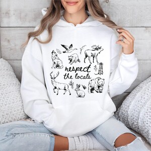 Puede incluir: Sudadera blanca con un gr&aacute;fico en blanco y negro de varios animales norteamericanos, incluyendo un lobo, un oso y un ciervo. La frase "respect the locals" est&aacute; impresa en un estilo manuscrito. La sudadera tiene un bolsillo delantero y una capucha con cord&oacute;n.
