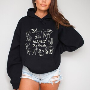 Puede incluir: Sudadera con capucha negra con un gr&aacute;fico color crema de varios animales, incluyendo un lobo, un mapache, un ciervo y un oso. El texto "respect the locals" est&aacute; centrado debajo de las ilustraciones de animales. La sudadera tiene una capucha con cord&oacute;n y un bolsillo tipo canguro.