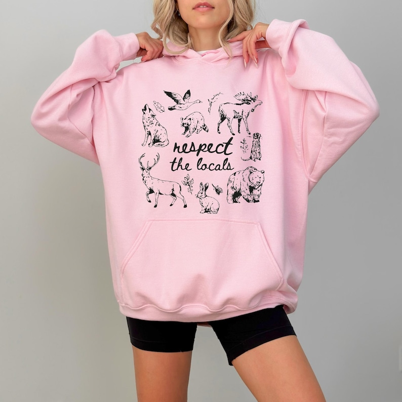 Puede incluir: Una sudadera rosa claro con bolsillo de canguro y capucha con cord&oacute;n. La parte delantera presenta un gr&aacute;fico negro de varios animales y el texto "respect the locals".