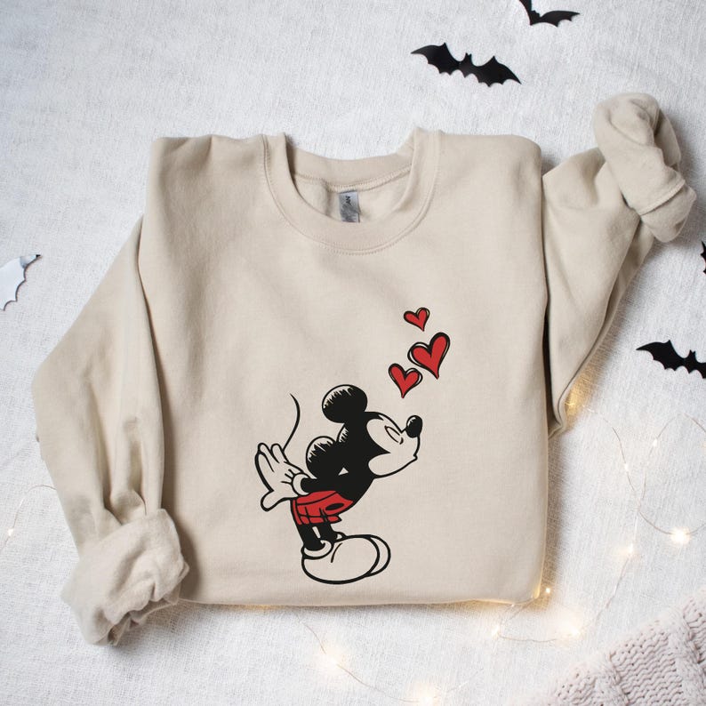 K&ouml;nnte beinhalten: Beiges Sweatshirt mit einem Cartoon-Design von Mickey Mouse, der rote Herzen pustet. Mickey ist in Schwarz, Rot und Wei&szlig; dargestellt. Das Sweatshirt hat einen Rundhalsausschnitt und lange &Auml;rmel. Dekorative Flederm&auml;use sind im Hintergrund.