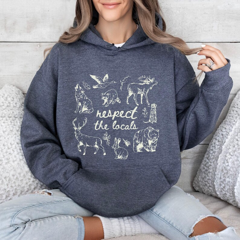 Puede incluir: Sudadera con capucha gris oscuro con un gr&aacute;fico blanco de varios animales, incluyendo un lobo, un alce y un oso, con el texto "respect the locals". La sudadera tiene un bolsillo delantero y una capucha con cord&oacute;n.