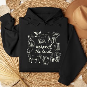Puede incluir: Sudadera con capucha negra con un gr&aacute;fico blanco de varios animales, incluyendo un lobo, un oso y un ciervo, con el texto "respect the locals". El dise&ntilde;o est&aacute; centrado en la parte delantera.