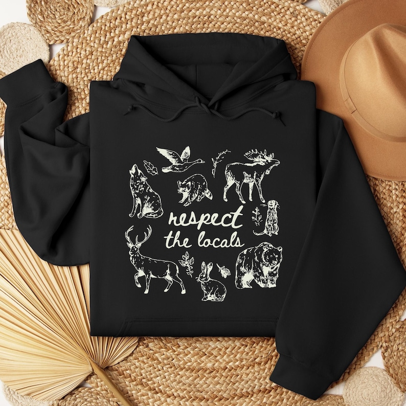 Puede incluir: Sudadera con capucha negra con un gr&aacute;fico blanco de varios animales, incluyendo un lobo, un oso y un ciervo, con el texto "respect the locals". El dise&ntilde;o est&aacute; centrado en la parte delantera.